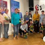 1. Grupa jedenastu uśmiechniętych ludzi, kobiet i mężczyzn. Stoją w grupie, przodem, część kuca, trzymają w rękach kolorowe, wielkanocne koszyki. W tle są kolorowe obrazy