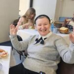 Uśmiechnięta kobieta, trzymająca ręce w górze, siedzi przy stole, w tle inna kobieta je śniadanie 