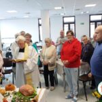 Grupa ludzi w różnym wieku, ksiądz stoi z otwartym Pismem Świętym, błogosławi pokarmy znajdujące się na stole, jajka, kiełbasę, baranka, mazurka