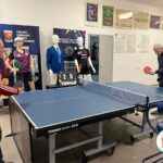 Dwóch mężczyzn gra w tenisa stołowego, w tle stoją manekiny w sportowych ubraniach, na ścianach wiszą różne plakaty